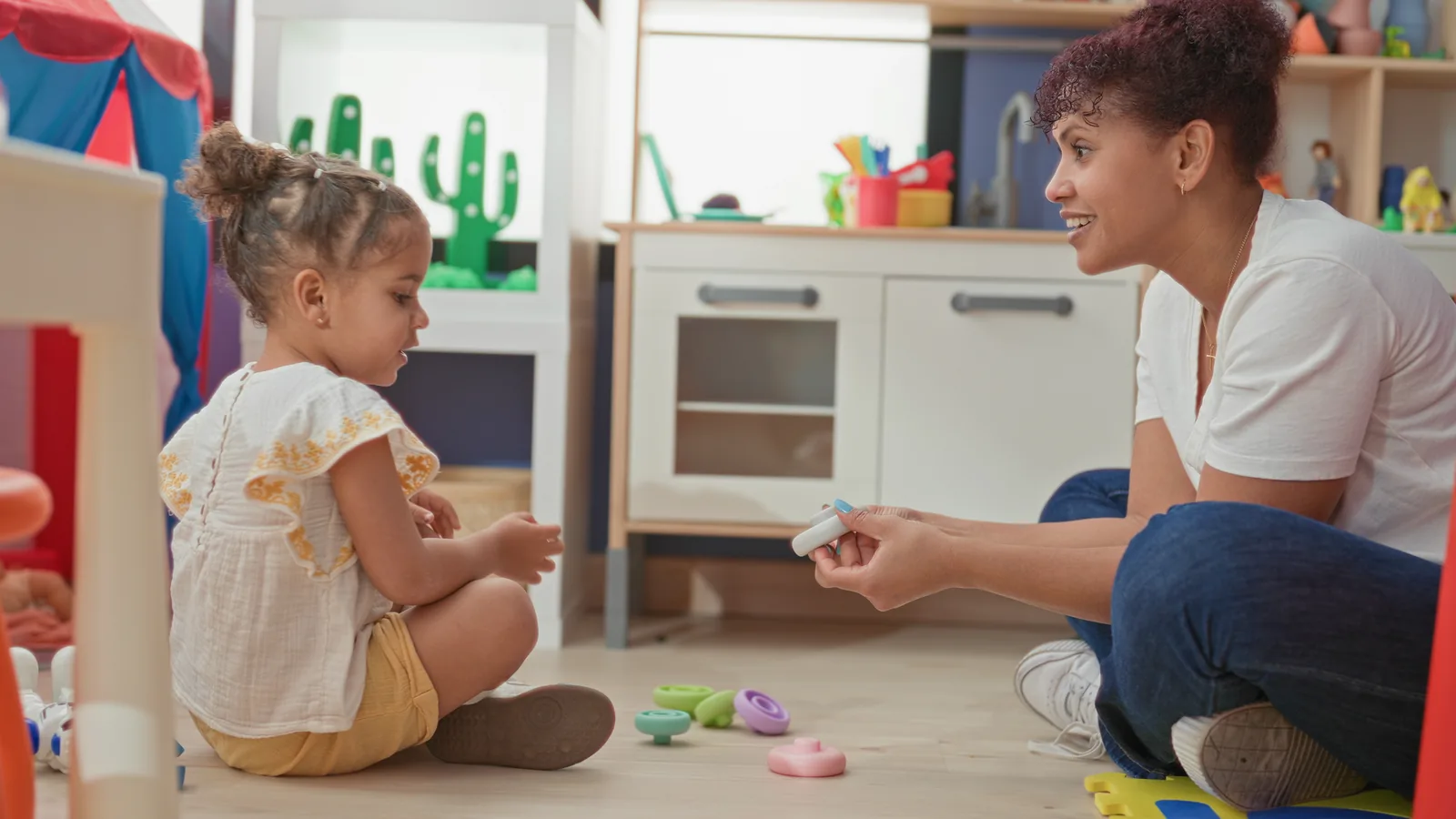A therapist crouches beside a young girl on the floor, handing her a toy in a playroom.
