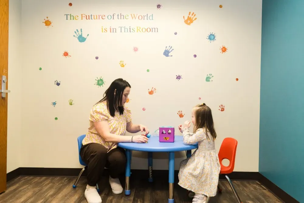 Un miembro del personal está sentado con un niño pequeño en una mesa pequeña dentro de un salón de terapia colorido del centro de Aurora, jugando juntos durante una sesión.