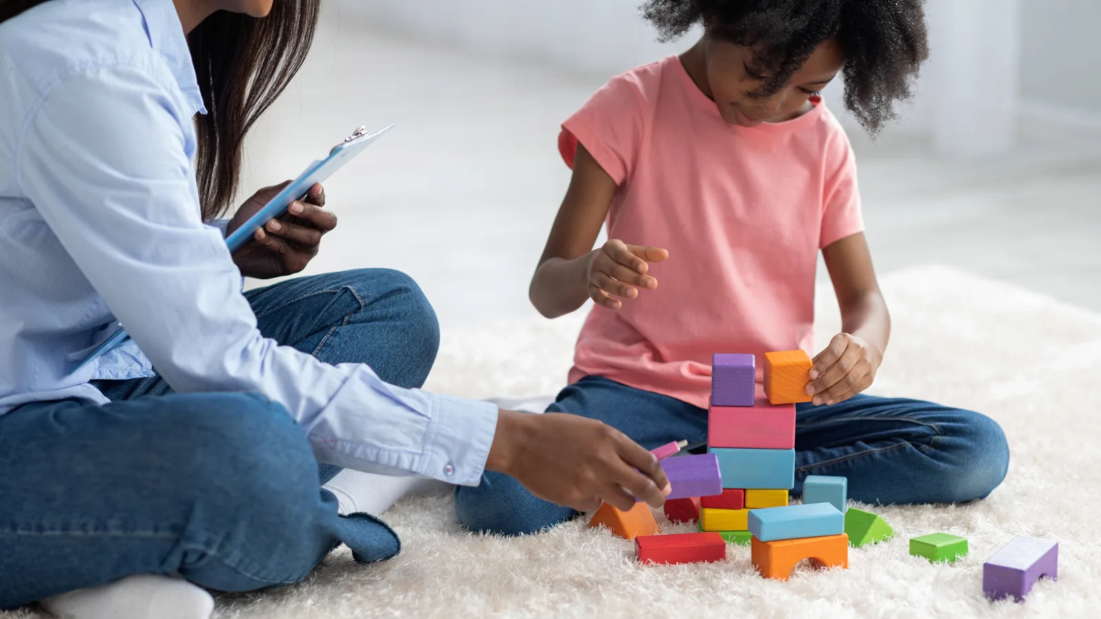 A therapist sits on the floor with a young girl, taking notes on a clipboard while she plays with wooden blocks.