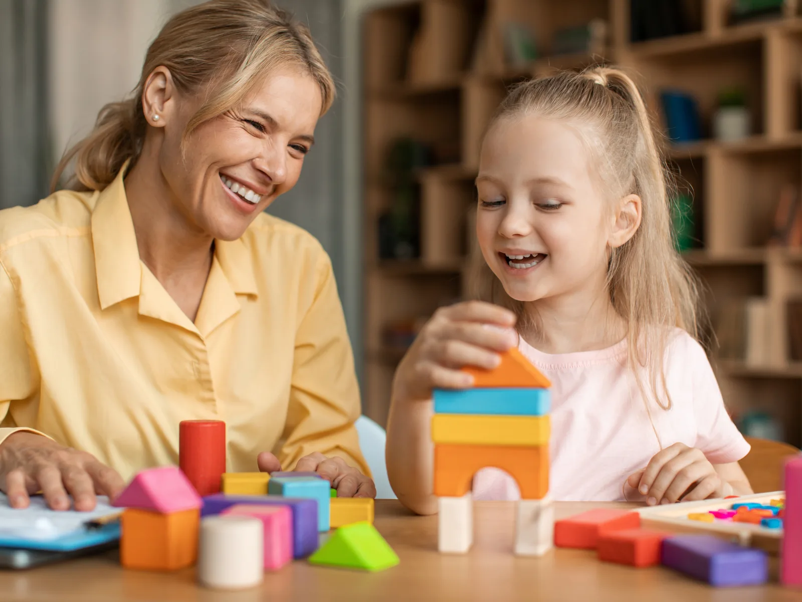 A therapist and a young girl smile while stacking colorful wooden blocks together at a table.