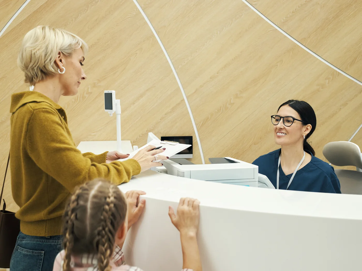 A parent checks in with a receptionist at the front desk while her daughter waits beside her.