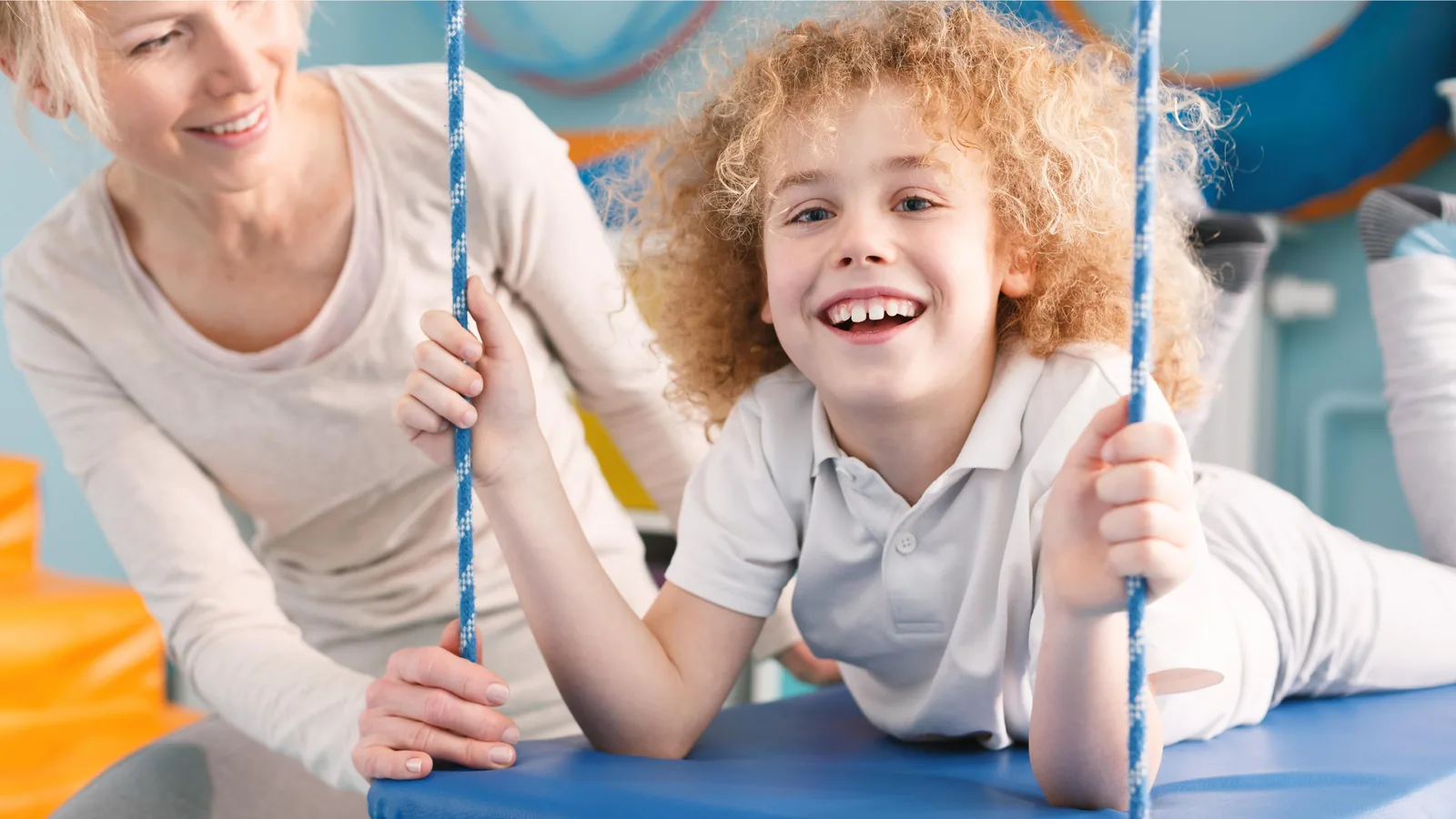 Un niño está recostado en un columpio de plataforma sonriendo a la cámara mientras una terapeuta sostiene las cuerdas detrás de él.