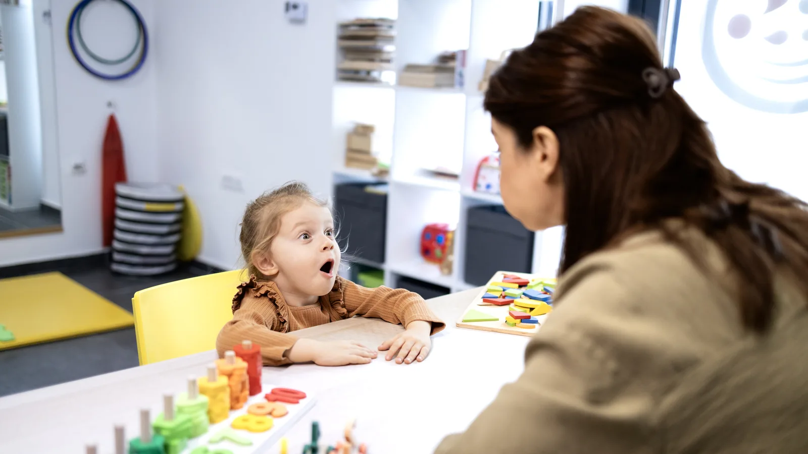 A young girl makes an exaggerated open-mouth expression at the table while a speech therapist practices sounds with her using colorful letter and number tiles.