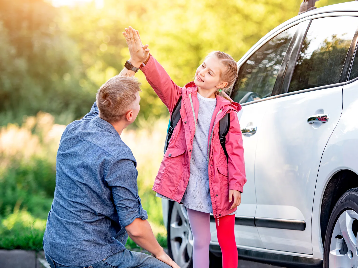 A father gives his daughter a high five outside a car as she arrives wearing a backpack.